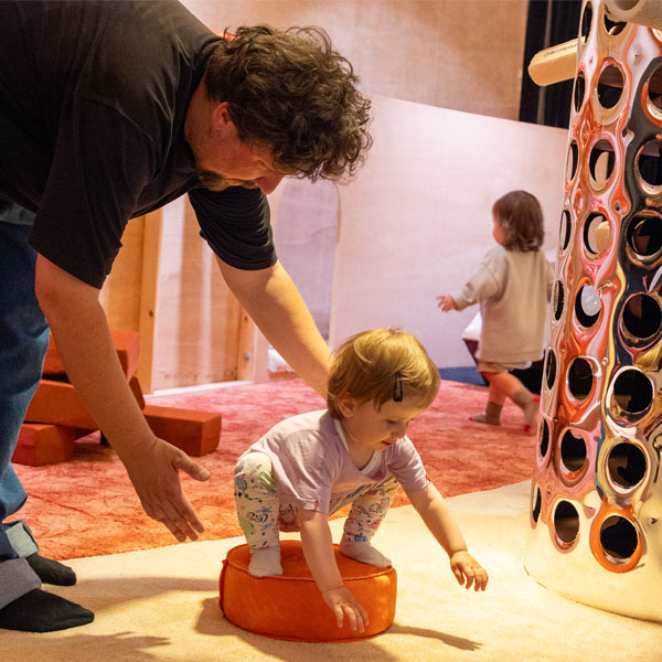 A man leans down to guide a toddler in a soft play area