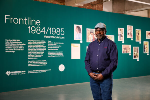 An older black man wearing a blue shirt and grey flatcap poses in front of exhibition interpretation.