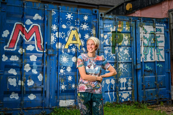 Artist, Peachezz, is stood in front of a blue container, with on hand on hip smiling to the side. The container has been painted in different colours with the words MAPA.