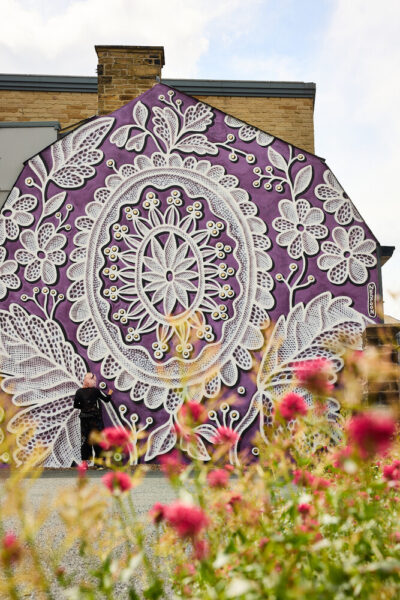 There is a blurred red flower bed that sits in the foreground of the image. Behind it is the mural, which depicts a lace pattern on a purple background. Artist, Nespoon is stood in the left hand corner of the building painting the wall.