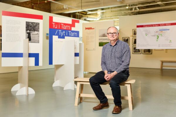 A white man with glasses sits in front of exhibition interpretation sat within the gallary.