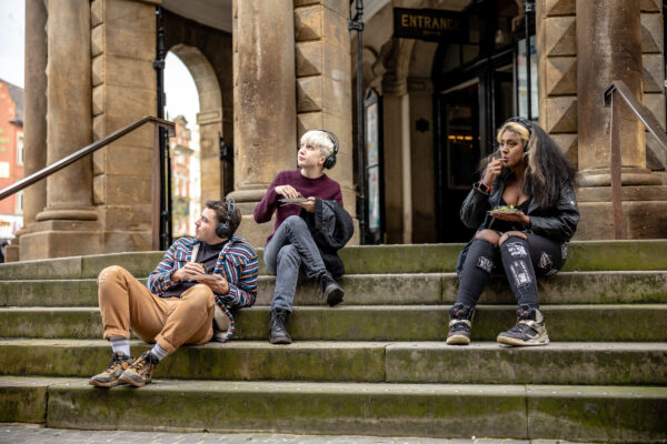 Three people sat on steps outside Kirkgate Market. Each have headphones on as they listen to the project's soundscape and eat the food prepared.