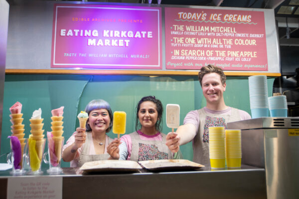 Three people stood behind an ice cream counter, holding an ice lolly / ice cream. There is a pink sign in the background which reads the ice cream menu.