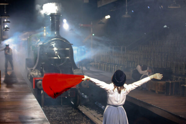 A girl stands on a train tracks in front of a large steam locomotive, waving a red hankerchief.