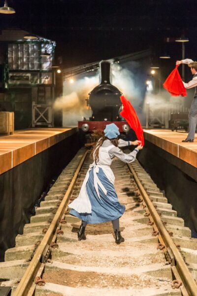 A girl stands on a train tracks in front of a large steam locomotive, waving a red hankerchief.