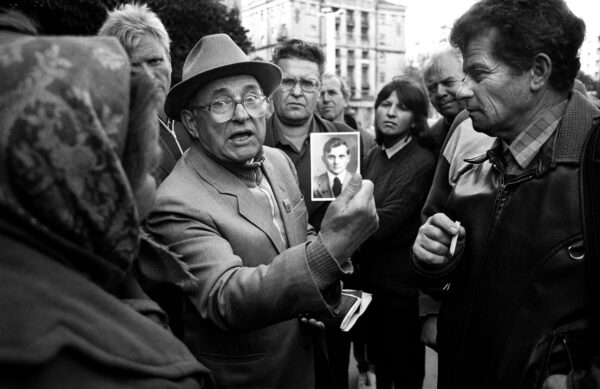 A black and white image. A crowd of people stand around a man in the centre of the image, holding a photograph of a boy.