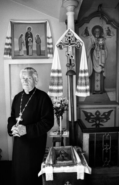 A black and white image. A priest wearing a large cross stands in front of religious artefacts.