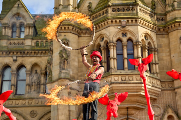 An acrobat in a red outfit similar to a traditional circus ringmaster holds flaming hoops as he is held aloft in front of several 8-meter tall red giraffe puppets, in front of Bradford City Hall.