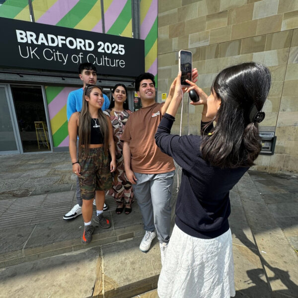 Four young people pose for a photo, as someone takes a photo of them on a mobile phone.