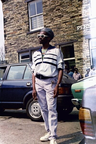 An archive photograph of a man wearing a white jacket and navy trousers. He is stood in front of a house and cars looking away from the camera into the distance.