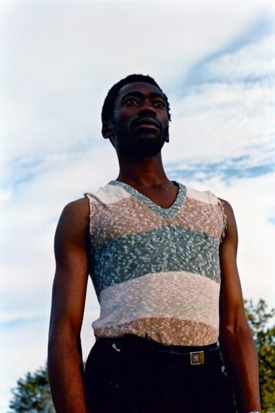 An archive photograph of a man wearing a blue, white and brown vest. He is looking over the camera into the distance.