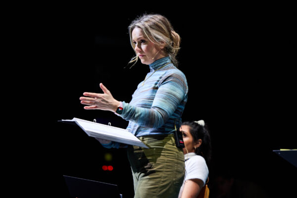 A woman with blonde hair and a blue jumper performs, reading from a black folder.