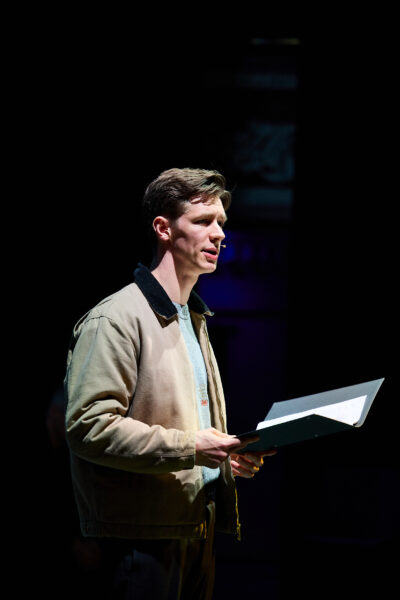 A man performs, reading from a black folder, downlit by stage lights.