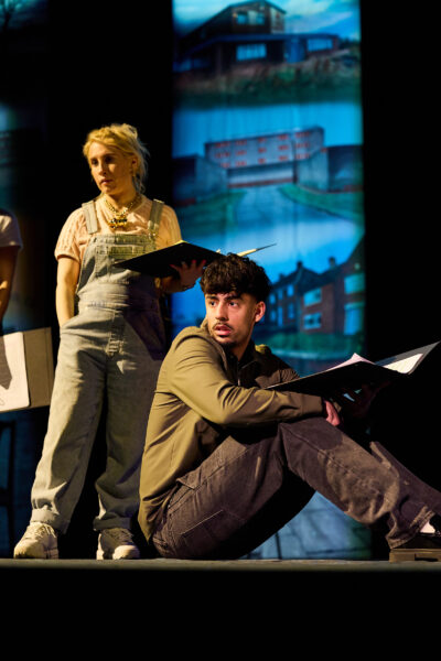 Two people perform on stage - a woman is stood and the man is sat.