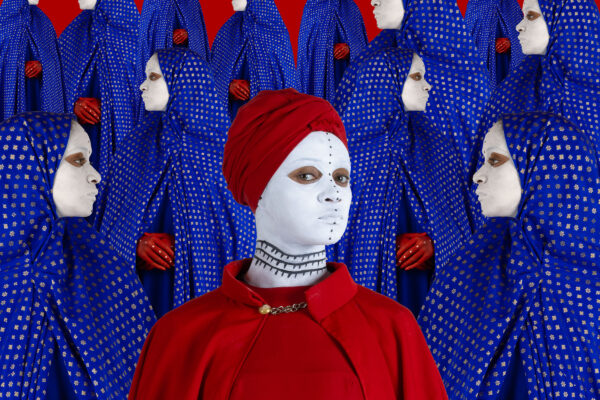 An image of a woman in a red cape and hijabi with a black and white painted face. She is surrounded by women in blue hijabis facing in different directions.