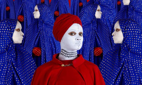 An image of a woman in a red cape and hijabi with a black and white painted face. She is surrounded by women in blue hijabis facing in different directions.