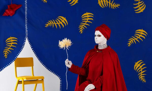 A woman with a white painted face in a red shawl and headscarf holds a white flower. She is sat in front of a blue background covered in yellow feathers. To the left of the image is a yellow wooden chair