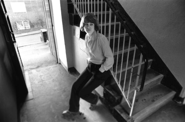 Andrea Dunbar sat leant against some stair railings. The image is in black and white and she is wearing long sleeve shirt and trousers.