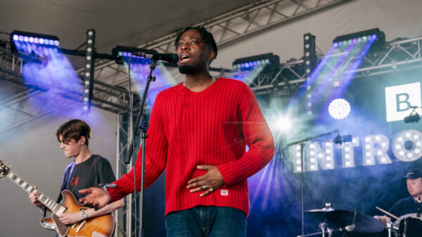 A band playing on stage for BBC Introducing. The lead singer is wearing a a red jumper and the bassist and drummer are in dark clothing in the background.