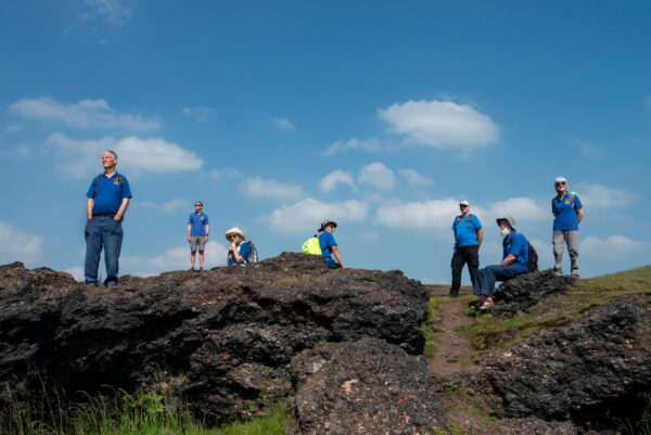 A group of walkers wear matching blue t-shirts. They are standing in a line on a rocky hill.