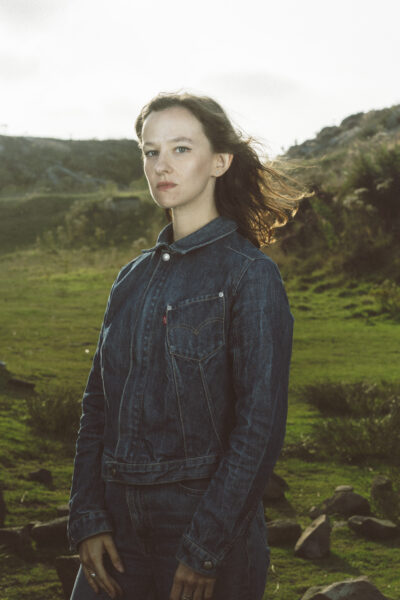 A woman with long dark hair and wearing a denim jack and jeans looks at the camera, there's a landscape behind her.