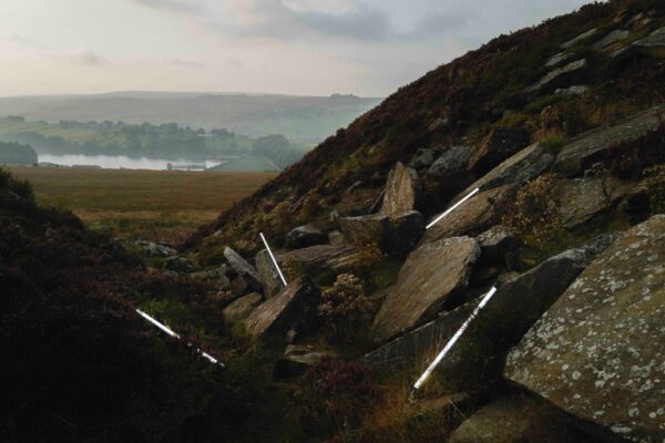 A dark moorland scene with rocks in the foreground and a reservoir in the background. Laid on the moorland are fluorescent lighting tubes.