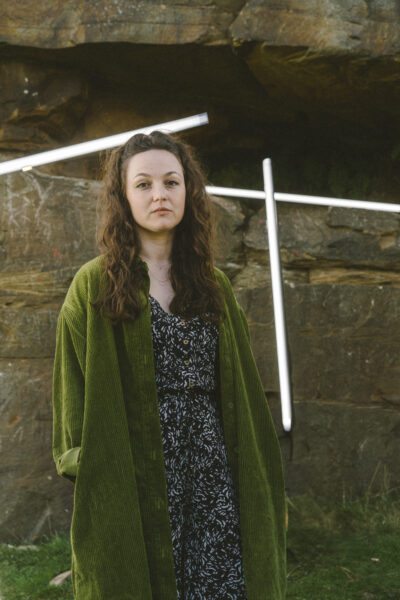 A woman wearing a patterned outfit and a long green jacket looks to the camera, there's a rock face with lights behind her.