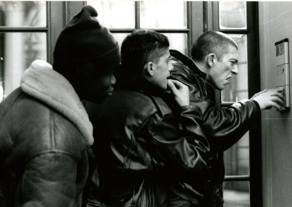 Three young men wearing jackets grouped together ring the doorbell for an apartment building. The picture is black and white.