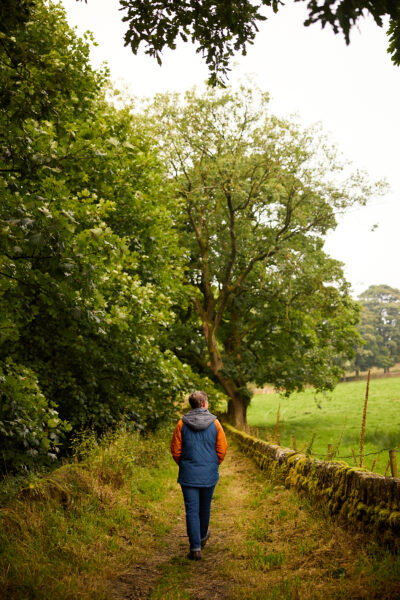 Person in blue gilet with hands in pockets walking away from the camera down a country path, a field is to the right of them.