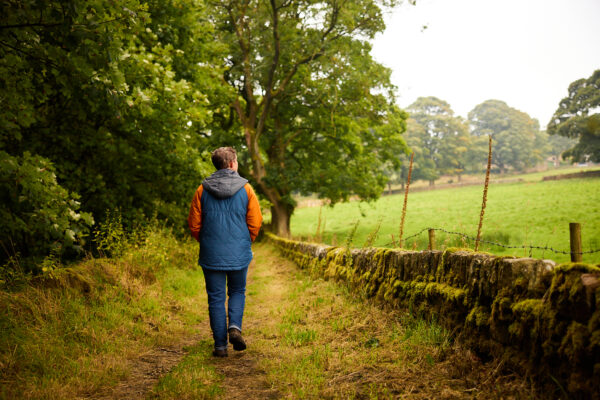 Person in blue gilet with hands in pockets walking away from the camera down a country path, a field is to the right of them.