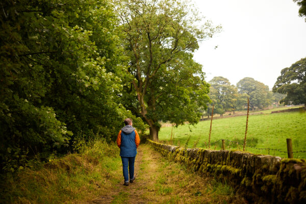 Person in blue gilet with hands in pockets walking away from the camera down a country path, a field is to the right of them.