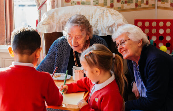 Two older ladies sit at a table with young children drawing