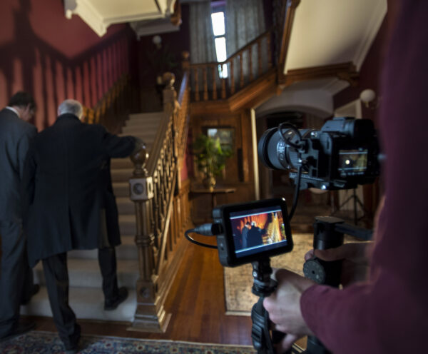 A behind the scenes image of a film shoot. The camera is in the foreground focussed on two people walking up some stairs.