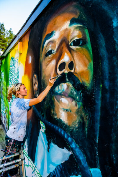 Large image of Mikey Roots, a Bradford community legend. Artist, Peachzz is stood in front of the image facing right with a paint brush and palette in her hand.