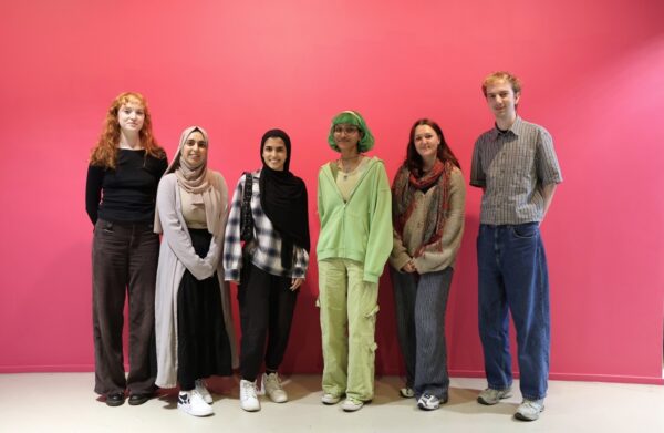 Six young people stand in a line, against a pink background.