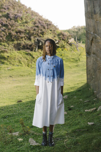 A woman wearing a blue and white dress looks at the camera, with a green landscape behind her.