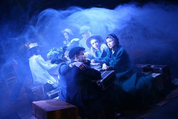 A group of actors sit on suitcases, they are all wearing Victorian era clothes and are surrounded by blue smoke.