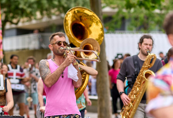 A man walking as part of a parade playing a brass instrument. He is wearing a pink vest and sunglasses.