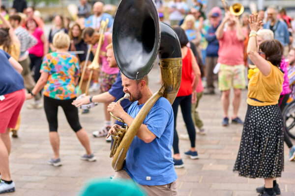 A man walking as part of a parade playing a brass instrument. He is wearing a blue t-shirt