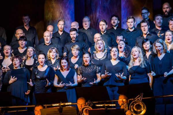 A chorus of singers all dressed in dark blue perform on a balcony above the orchestra.