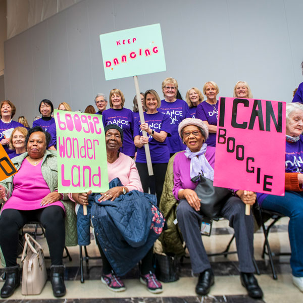 A group of older ladies sit together holding up placards with phrases including 'I Can Boogie' and Boogie Wonderland' on them.