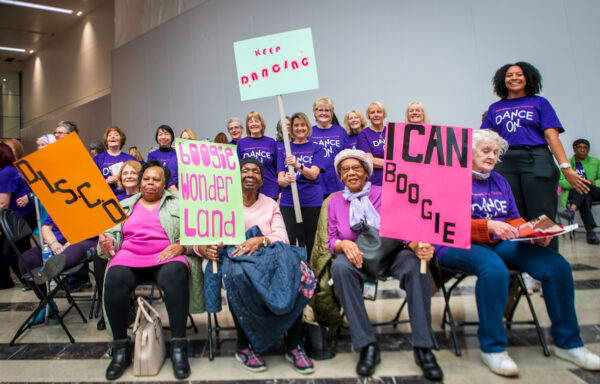 A group of older ladies sit together holding up placards with phrases including 'I Can Boogie' and Boogie Wonderland' on them.