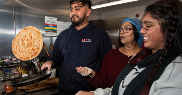 Three individuals standing over a cooking hob, where two of them are flipping chapatis in the pan. One is being tossed in the air.