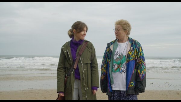 Two people smile at each other on a beach, with a grey sky behind. A still from the film Typist Artist Pirate King.