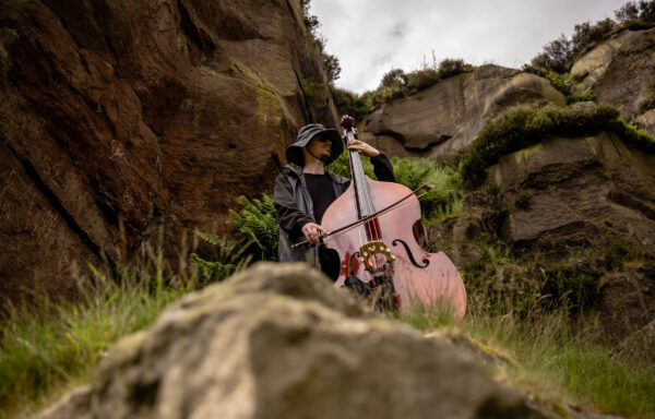 A man in a black mac and black rain hat plays a double bass on some rocks in the countryside.