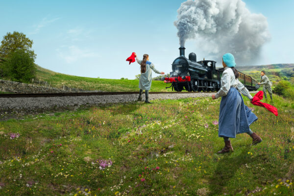 Three children in Victorian dress wave red flags at a steam train, on a sunny day in West Yorkshire.