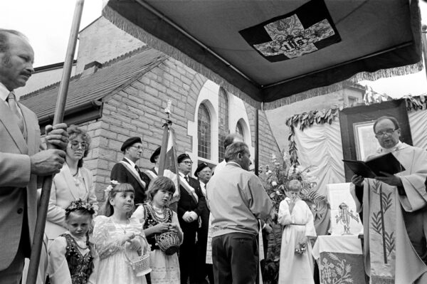 A group of adults and children stood a priest who is reading from a book.