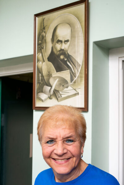 A smiling woman stands below a framed image of a man. Their faces both look towards us.