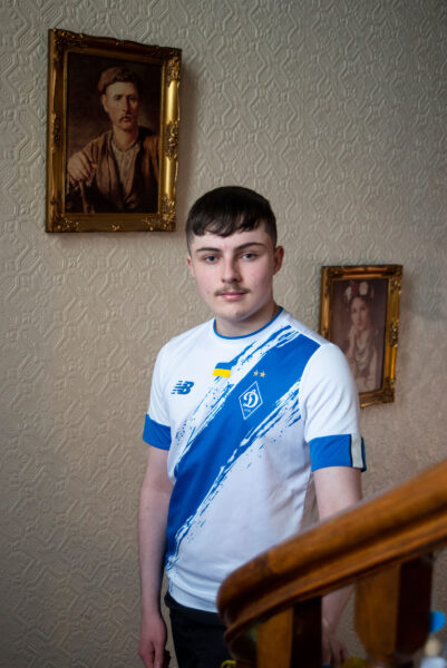 A young man wearing a Ukrainian sports shirt stands in front of framed pictures on a wall.