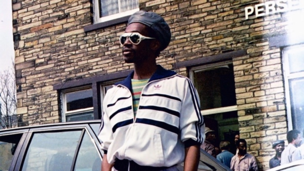 Man with white sunglasses on is looking to his left, he is wearing a black and white striped sports top. He is stood in front of a bricked building and a car